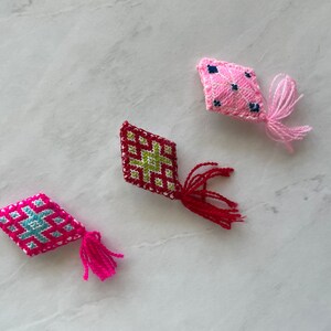 May include: Three embroidered diamond-shaped ornaments with tassels. The ornaments are pink, red, and green with white accents.