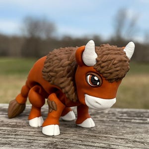 May include: A brown and orange bison figurine with white horns and hooves. The bison has a textured brown mane and tail. The figurine is posed on a wooden surface with a blurred background of trees and sky.