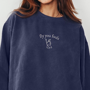 May include: A navy blue sweatshirt with the embroidered phrase "fly you fools" and a wizard graphic. The long-sleeved crew neck top is made of a soft material and is suitable for casual wear.