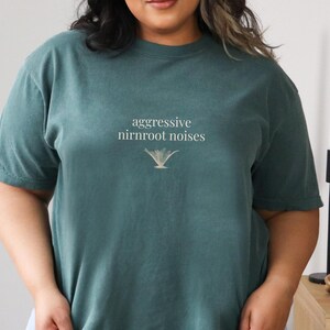 May include: A teal-colored t-shirt with the text "aggressive nirnroot noises" in a white, stylized font. Below the text is a gold-colored graphic. The shirt has short sleeves and a crew neck.