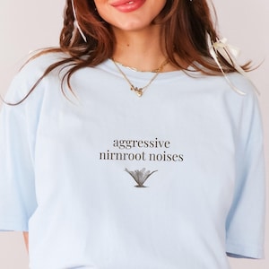 May include: Light blue t-shirt with the text "aggressive nirnroot noises" and a small illustration of a plant. The shirt is a soft, casual style.