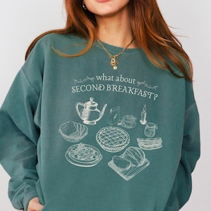 May include: Teal-colored sweatshirt with the text "what about SECOND BREAKFAST?" and a white illustration of breakfast items. The graphic includes a teapot, bread, pie, and other food items. The sweatshirt is a relaxed fit.