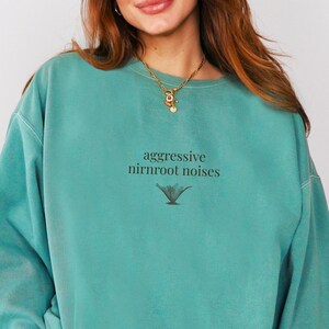 May include: Teal-colored sweatshirt with the text "aggressive nirnroot noises" in black, along with a small graphic. The sweatshirt has a crew neck and long sleeves. The fabric appears soft and comfortable.