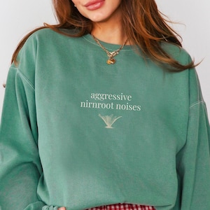 May include: A teal green sweatshirt with the text "aggressive nirnroot noises" in white, and a small graphic below. The sweatshirt has long sleeves and a relaxed fit. The bottom of the sweatshirt is not visible.