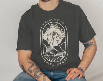 May include: A dark gray t-shirt with a white graphic design. The graphic features a mountain range with a staircase leading up to a cave. The text "Welcome to Cirith Ungol" is written in a circle around the graphic.