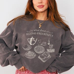 May include: Dark gray sweatshirt with the text "what about SECOND BREAKFAST?" and a detailed illustration of breakfast foods. The design includes a teapot, pie, and other breakfast items. The sweatshirt has long sleeves.