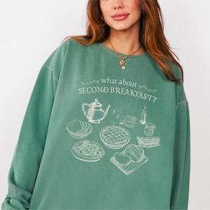 May include: A teal green sweatshirt with the phrase "what about SECOND BREAKFAST?" and a graphic of breakfast foods. The graphic includes a teapot, pie, and other breakfast items. The sweatshirt is a relaxed fit.