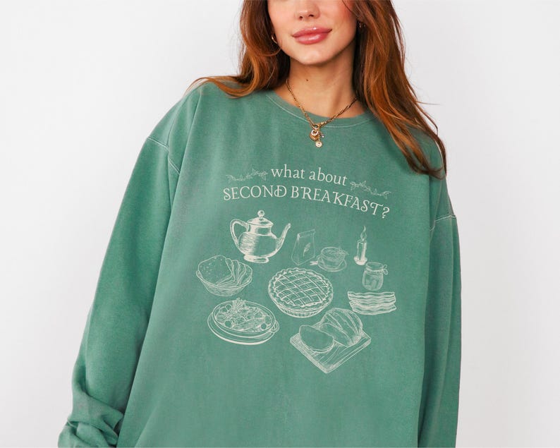 May include: A teal green sweatshirt with the phrase "what about SECOND BREAKFAST?" and a graphic of breakfast foods. The graphic includes a teapot, pie, and other breakfast items. The sweatshirt is a relaxed fit.
