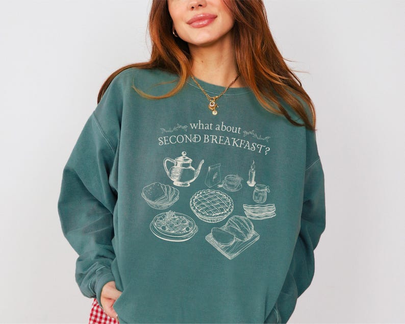 May include: Teal-colored sweatshirt with the text "what about SECOND BREAKFAST?" and a white illustration of breakfast items. The graphic includes a teapot, bread, pie, and other food items. The sweatshirt is a relaxed fit.