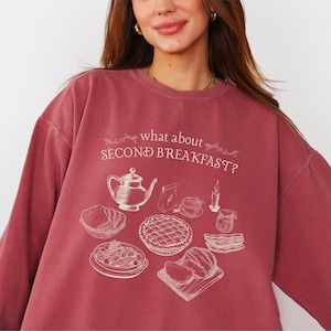 May include: A maroon sweatshirt with the text "what about SECOND BREAKFAST?" and a cream-colored illustration of breakfast foods. The illustration includes a teapot, pie, bacon, and other breakfast items. The sweatshirt has long sleeves.
