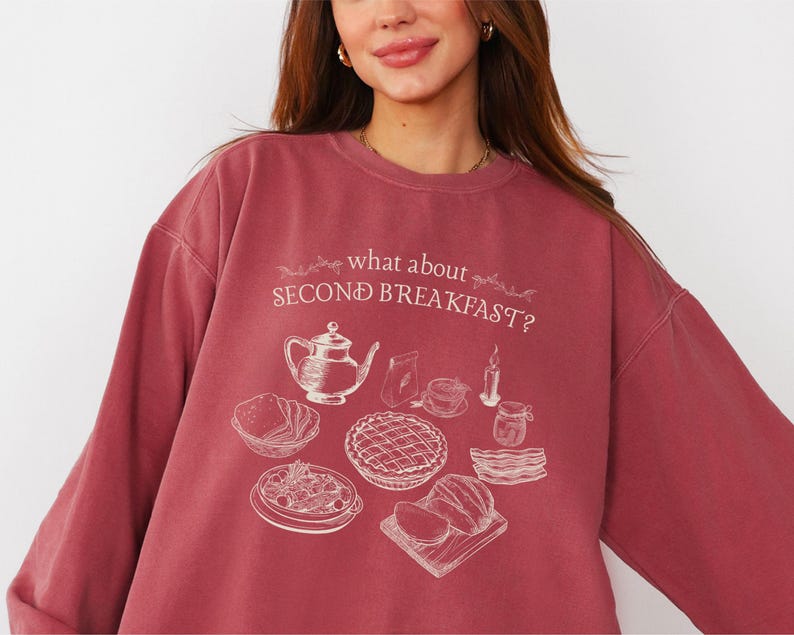 May include: A maroon sweatshirt with the text "what about SECOND BREAKFAST?" and a cream-colored illustration of breakfast foods. The illustration includes a teapot, pie, bacon, and other breakfast items. The sweatshirt has long sleeves.