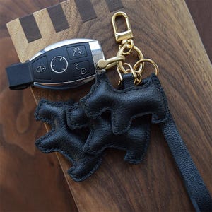 K&ouml;nnte beinhalten: Schwarzer Leder-Schl&uuml;sselanh&auml;nger in Hundeform mit goldfarbenem Verschluss und Schl&uuml;sselring. Der Schl&uuml;sselanh&auml;nger ist an einem schwarzen Lederband befestigt. Ein Autoschl&uuml;ssel ist ebenfalls sichtbar.