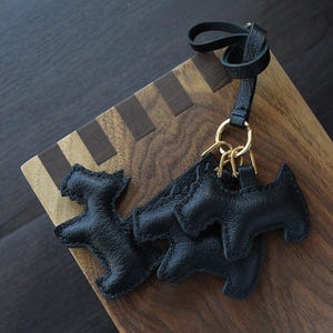 K&ouml;nnte beinhalten: Schwarze Leder-Schl&uuml;sselanh&auml;nger in Hundeform mit goldfarbener Hardware. Die Schl&uuml;sselanh&auml;nger sind an einem schwarzen Lederband befestigt. Die Schl&uuml;sselanh&auml;nger liegen auf einer Holzoberfl&auml;che mit einem dunkelbraunen und hellbraunen Muster.