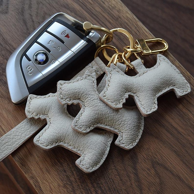 K&ouml;nnte beinhalten: Ein silberner Autoschl&uuml;ssel und ein Satz von drei hellgrauen Leder-Schl&uuml;sselanh&auml;ngern in Hundeform mit goldfarbener Hardware. Die Schl&uuml;sselanh&auml;nger liegen auf einer Holzoberfl&auml;che.