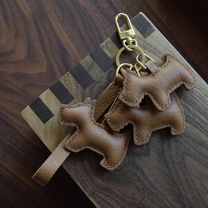 K&ouml;nnte beinhalten: Braune Leder-Schl&uuml;sselanh&auml;nger in Hundeform mit goldfarbener Hardware. Die Schl&uuml;sselanh&auml;nger sind an einem goldenen Verschluss und einem Lederriemen befestigt. Die Schl&uuml;sselanh&auml;nger sind aus strukturiertem braunem Leder gefertigt und an den R&auml;ndern vern&auml;ht. Der Hintergrund ist eine Holzoberfl&auml;che.