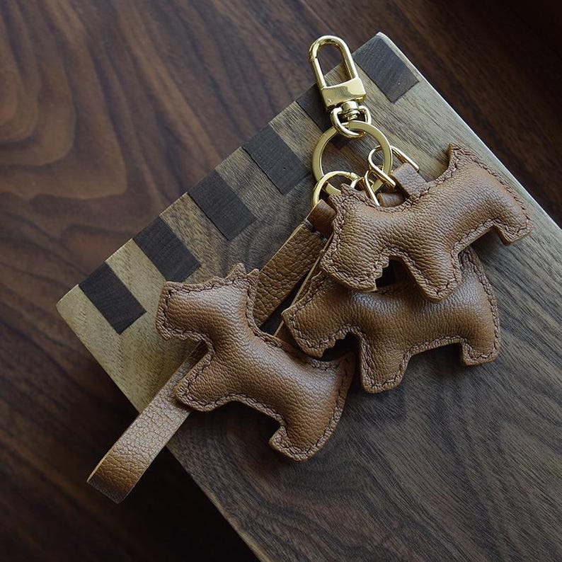 K&ouml;nnte beinhalten: Braune Leder-Schl&uuml;sselanh&auml;nger in Hundeform mit goldfarbener Hardware. Die Schl&uuml;sselanh&auml;nger sind an einem goldenen Verschluss und einem Lederriemen befestigt. Die Schl&uuml;sselanh&auml;nger sind aus strukturiertem braunem Leder gefertigt und an den R&auml;ndern vern&auml;ht. Der Hintergrund ist eine Holzoberfl&auml;che.