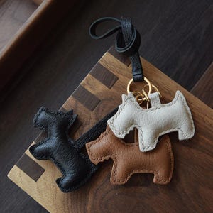 K&ouml;nnte beinhalten: Drei lederne Schl&uuml;sselanh&auml;nger in Hundeform in Schwarz, Hellbraun und Off-White. Die Schl&uuml;sselanh&auml;nger sind an einem schwarzen Lederband und einem goldfarbenen Metallring befestigt. Die Schl&uuml;sselanh&auml;nger liegen auf einer Holzoberfl&auml;che.