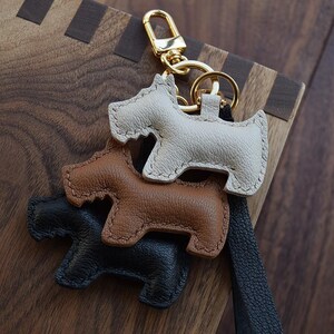 K&ouml;nnte beinhalten: Drei lederne Schl&uuml;sselanh&auml;nger in Hundeform in Creme, Braun und Schwarz. Jeder Schl&uuml;sselanh&auml;nger hat eine gen&auml;hte Kontur und einen goldfarbenen Metallverschluss. Die Schl&uuml;sselanh&auml;nger sind &uuml;bereinander gestapelt, mit einem schwarzen Lederriemen.