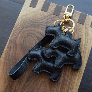 K&ouml;nnte beinhalten: Schwarzer Leder-Schl&uuml;sselanh&auml;nger mit drei stilisierten Hundesilhouetten. Er hat einen goldfarbenen Verschluss und Schl&uuml;sselring sowie einen schwarzen Lederriemen. Die Hundeformen sind mit schwarzem Faden gen&auml;ht. Der Hintergrund ist eine Holzoberfl&auml;che.