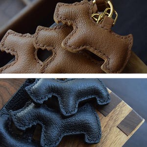 K&ouml;nnte beinhalten: Nahaufnahme von zwei ledernen Schl&uuml;sselanh&auml;ngern in Hundeform. Einer ist braun mit einem goldfarbenen Verschluss, der andere ist schwarz. Die Schl&uuml;sselanh&auml;nger sind mit einem doppelten Sattelstich gefertigt. Der Text "Handmade Double-Needle Saddle Stitch" und "French Imported Top-Grain Leather" sind sichtbar.