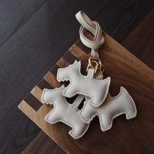 K&ouml;nnte beinhalten: Hellwei&szlig;e Leder-Schl&uuml;sselanh&auml;nger in Form von Scottish Terrier-Hunden. Die Schl&uuml;sselanh&auml;nger sind an einer hellfarbigen Lederschlaufe mit einem goldfarbenen Metallverschluss befestigt. Auf einer Holzoberfl&auml;che.