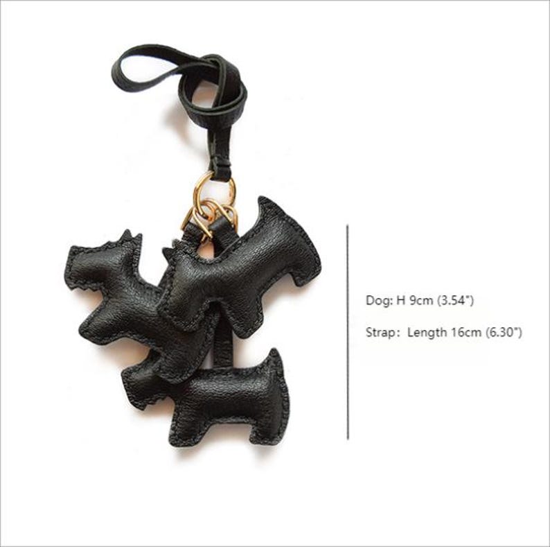 K&ouml;nnte beinhalten: Schwarzer Leder-Schl&uuml;sselanh&auml;nger in Hundeform mit goldenem Ring und schwarzem Riemen. Der Schl&uuml;sselanh&auml;nger zeigt drei Hundesilhouetten, jede etwa 9 cm hoch. Der Riemen ist etwa 16 cm lang.