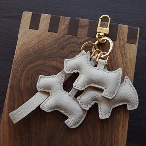 K&ouml;nnte beinhalten: Eine Sammlung hellgrauer Leder-Schl&uuml;sselanh&auml;nger in Hundeform mit goldfarbener Hardware. Die Schl&uuml;sselanh&auml;nger sind an einem goldfarbenen Karabiner und Ringen befestigt. Der Hintergrund ist eine Holzoberfl&auml;che mit dunkler brauner Oberfl&auml;che.