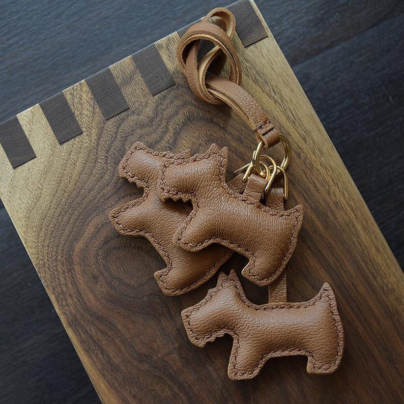 K&ouml;nnte beinhalten: Drei braune Leder-Schl&uuml;sselanh&auml;nger in Hundeform mit goldfarbener Hardware. Die Schl&uuml;sselanh&auml;nger sind an einer Lederschlaufe befestigt. Die Schl&uuml;sselanh&auml;nger liegen auf einer Holzoberfl&auml;che mit dunklem Maserungsmuster.