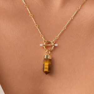 May include: A gold-toned necklace featuring a delicate chain with cylindrical links and a toggle clasp closure. The toggle bar is accented with two small clear stones. A polished, hexagonal tiger's eye gemstone pendant hangs from the centre ring.