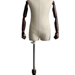 May include: A child-sized, beige fabric torso mannequin with wooden arms and a wooden base. The mannequin is on a metal stand with a wooden base.