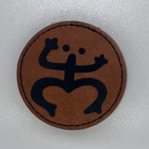 Puede incluir: Un parche de cuero marrón con un diseño de rana estilizado en negro. La rana está en posición sentada con los brazos levantados y las piernas extendidas.