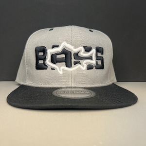Puede incluir: Una gorra de béisbol gris y negra con una silueta de pez blanca y la palabra "BASS" bordada en la parte delantera.