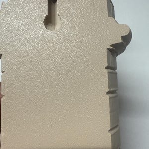 May include: A beige wall-mounted decorative item with a textured surface. The item has an arched top and decorative side elements. A keyhole-shaped opening is visible on the back for hanging.