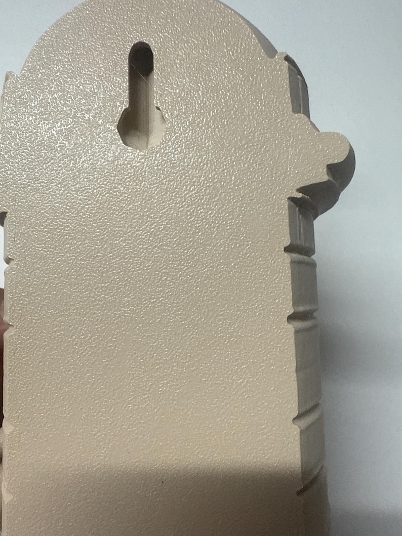 May include: A beige wall-mounted decorative item with a textured surface. The item has an arched top and decorative side elements. A keyhole-shaped opening is visible on the back for hanging.