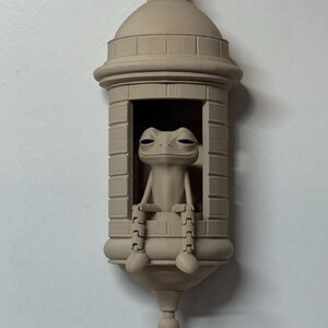 May include: A beige-colored decorative frog figurine seated inside a small, architectural structure. The frog has a relaxed expression, with its legs and feet visible. The structure has a domed roof and a decorative base.