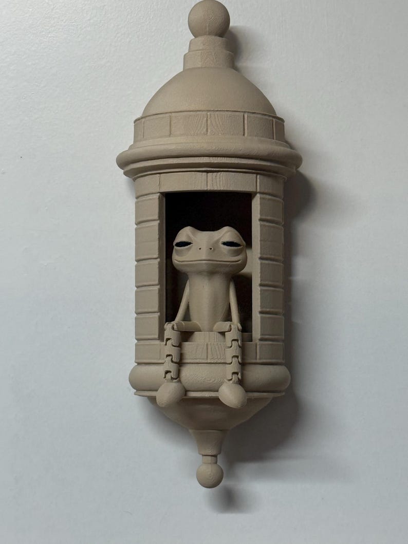 May include: A beige-colored decorative frog figurine seated inside a small, architectural structure. The frog has a relaxed expression, with its legs and feet visible. The structure has a domed roof and a decorative base.