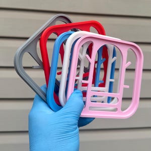 May include: A stack of five square, open-frame coasters in gray, red, blue, white, and pink. Each coaster features a stylized tree branch design integrated into the frame, with a smooth, matte finish and rounded corners on the outer edges.