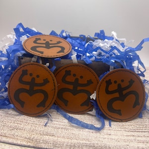 May include: Four brown leather patches with a black stylized symbol of a frog or a person with arms raised. The patches are in a blue and white shredded paper gift box.