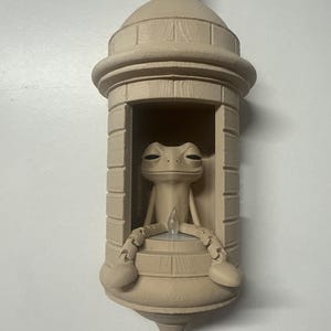 May include: A beige-colored wall decoration featuring a frog figurine seated inside a miniature tower. The frog has its arms and legs resting on the tower's ledge. The tower has a domed roof and a decorative base.