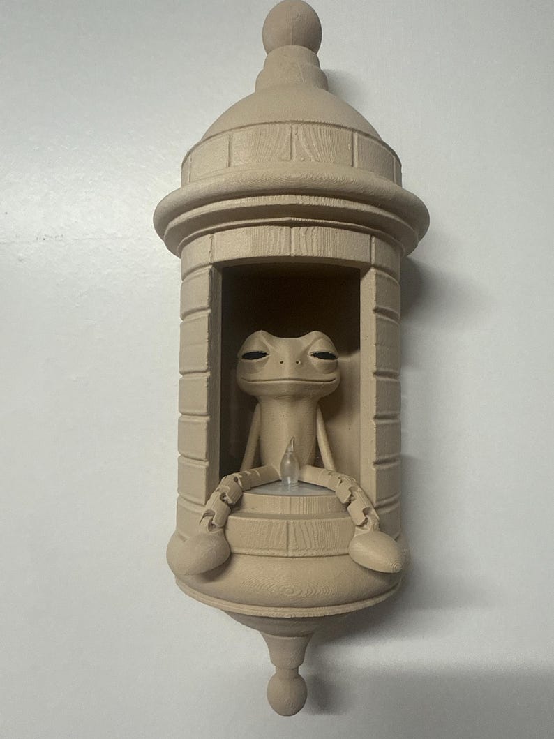 May include: A beige-colored wall decoration featuring a frog figurine seated inside a miniature tower. The frog has its arms and legs resting on the tower's ledge. The tower has a domed roof and a decorative base.