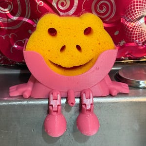 May include: A yellow frog-shaped sponge sits in a pink holder with a smiling face. The holder has pink legs and arms, designed to sit on a sink. The sponge has two eye holes and a smiling mouth.