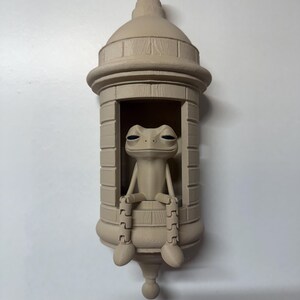 May include: A beige-colored frog figurine seated inside a decorative, cylindrical structure. The frog has its legs dangling over the edge. The structure has a domed top and a rounded base, resembling a bottle or a small tower.