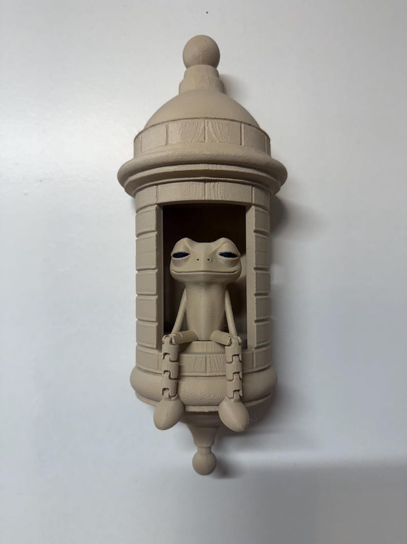 May include: A beige-colored frog figurine seated inside a decorative, cylindrical structure. The frog has its legs dangling over the edge. The structure has a domed top and a rounded base, resembling a bottle or a small tower.