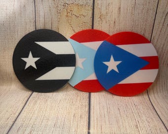 May include: Three round coasters featuring the flag of Puerto Rico. The coasters are black, red, and blue, with white stars and stripes. They are arranged on a light brown wooden surface.