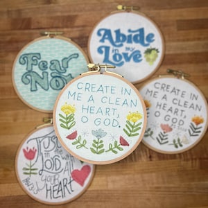 May include: Five embroidered hoop art pieces with inspirational quotes. The hoops feature text like "Fear No" and "Abide in my Love." Floral and heart designs accompany the text, with a wooden background.