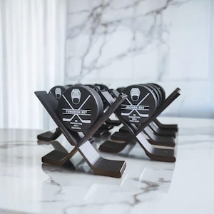 May include: A set of black hockey puck awards with personalised names and numbers on each puck. The pucks are displayed on a black wooden stand shaped like hockey sticks.