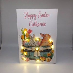 May include: Easter greeting card with the text "Happy Easter Catherine". The card features a painted illustration of two bunnies in a wooden basket with Easter eggs, flowers, and a chick. The card is illuminated with small, warm white lights.