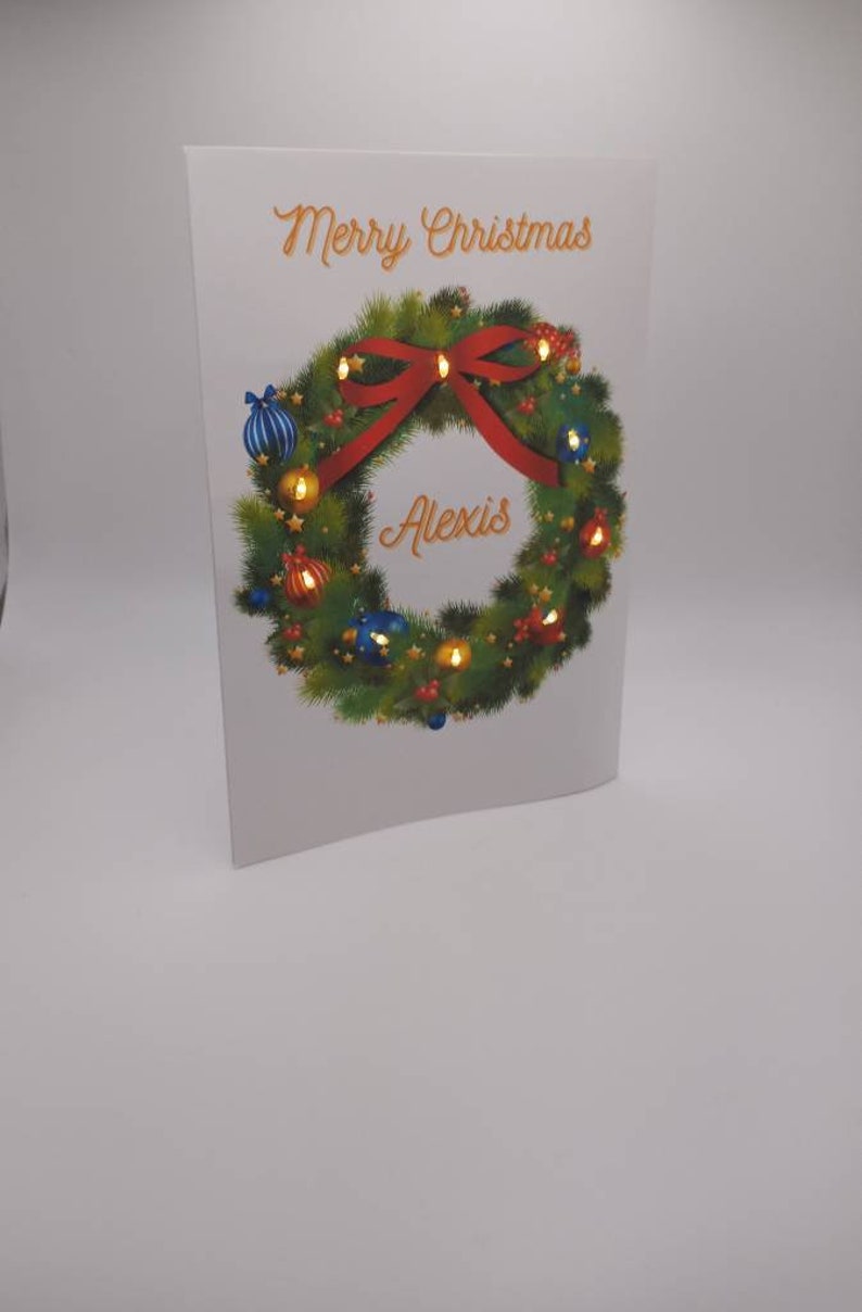 K&ouml;nnte beinhalten: Eine Weihnachtskarte mit einem festlichen Kranzdesign. Der Kranz besteht aus gr&uuml;nen Tannennadeln, verziert mit roten und blauen Kugeln und einer roten Schleife. Die Worte "Merry Christmas" und der Name "Alexis" sind auf der Karte geschrieben.