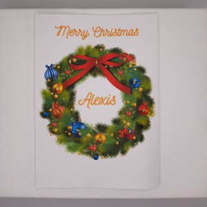 K&ouml;nnte beinhalten: Eine Weihnachtskarte mit einem Kranzdesign. Der Kranz besteht aus gr&uuml;nen Tannennadeln, verziert mit roten, blauen und goldenen Kugeln und einer roten Schleife. Die Worte "Merry Christmas" und der Name "Alexis" sind in goldener Schrift geschrieben.