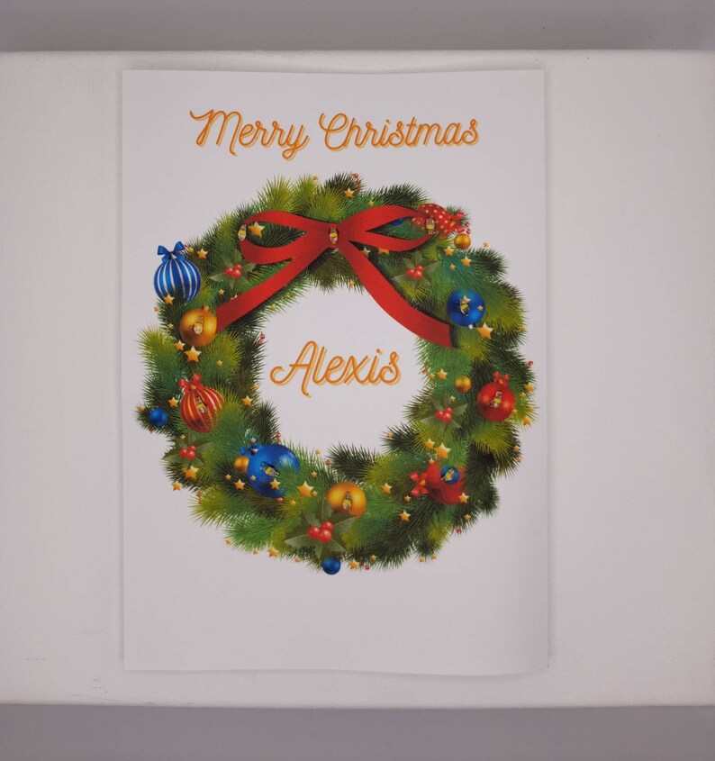 K&ouml;nnte beinhalten: Eine Weihnachtskarte mit einem Kranzdesign. Der Kranz besteht aus gr&uuml;nen Tannennadeln, verziert mit roten, blauen und goldenen Kugeln und einer roten Schleife. Die Worte "Merry Christmas" und der Name "Alexis" sind in goldener Schrift geschrieben.